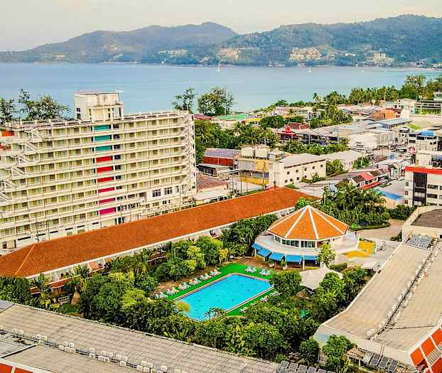 Utsikt over et hotellområde med en hvit fleretasjes bygning, et svømmebasseng og havutsikt. (AI-generert tekst)