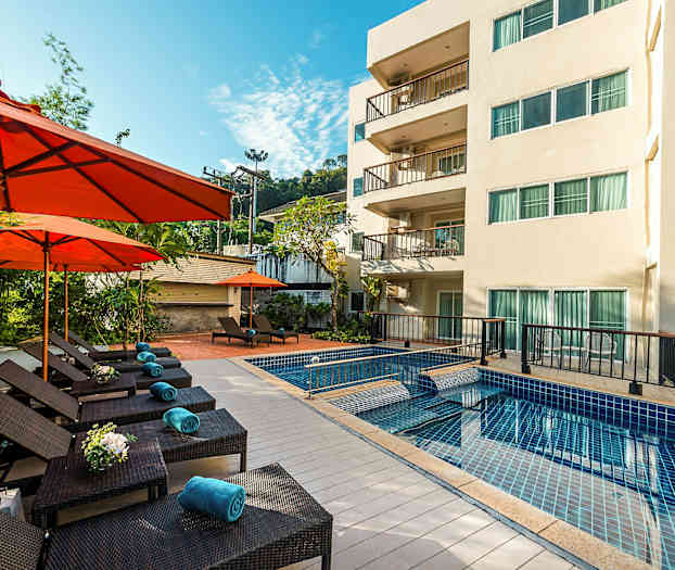 Utendørs basseng med solsenger og oransje parasoller ved siden av en treetasjes bygning. (AI-generert tekst)