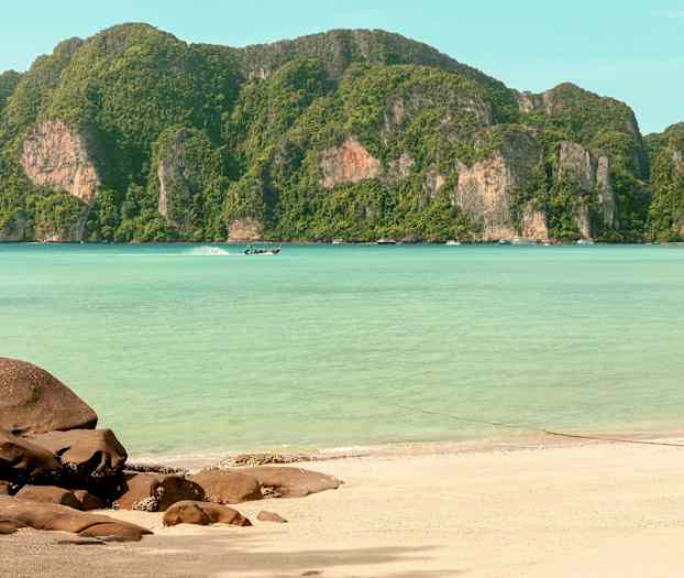 En sandstrand med steiner i forgrunnen, klart turkist vann og grønne fjell i bakgrunnen. (AI-generert tekst)