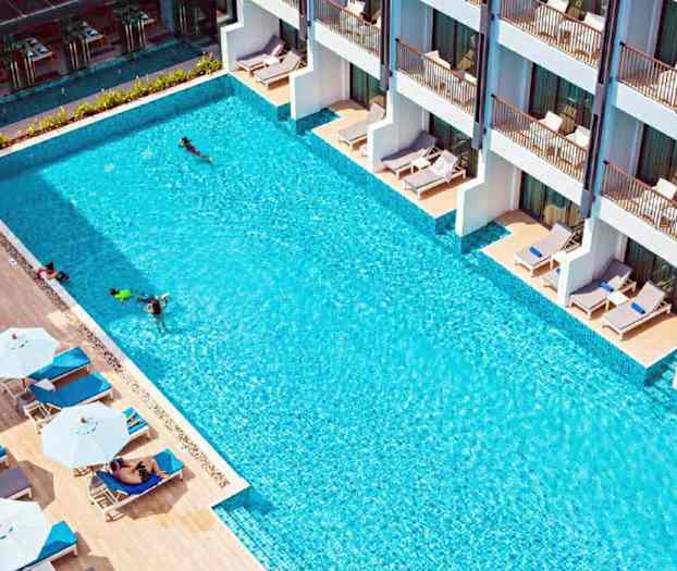 Et utendørs basseng med solsenger og parasoller på siden, samt bygninger med terrasser. (AI-generert tekst)