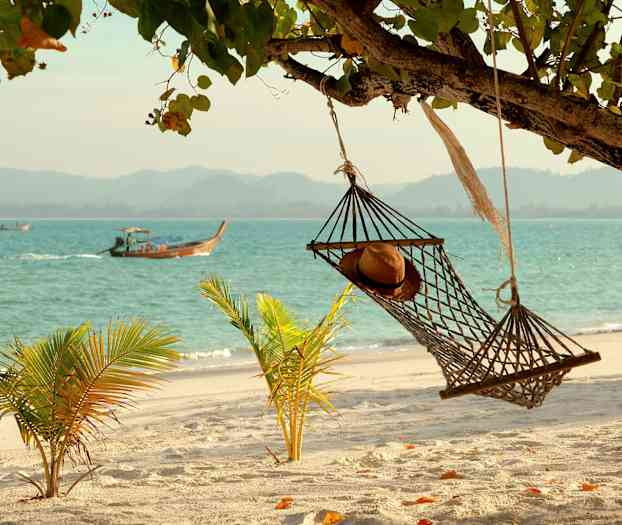 Hengekøye med en hatt ved en sandstrand, sjøen og båter i bakgrunnen. (AI-generert tekst)