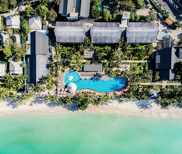Flyfoto av et feriested med basseng, omgitt av tropiske trær og nær en strand med klart vann. (AI-generert tekst)