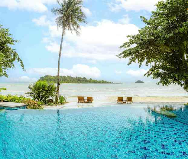 En utsikt over et utendørsbasseng i forgrunnen med en strand og havet i bakgrunnen, omgitt av frodige planter og palmer. (AI-generert tekst)