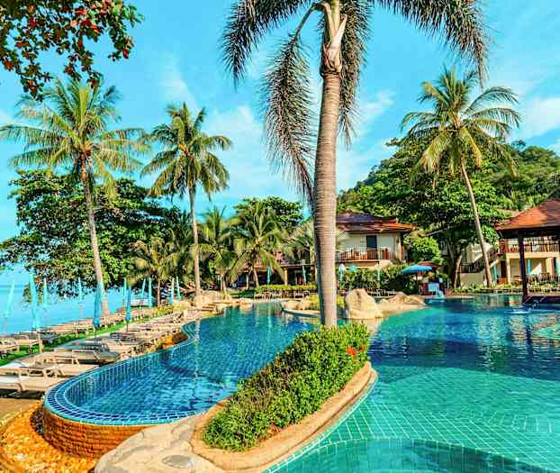 Et basseng med solsenger og parasoller omgitt av palmer ved siden av havet. (AI-generert tekst)