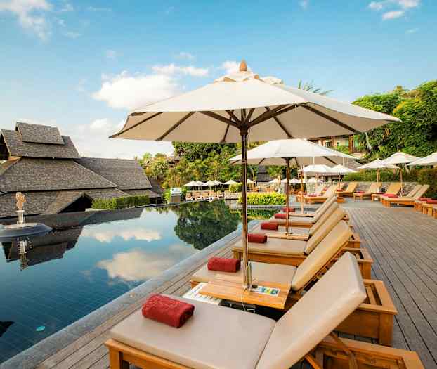 Bassengområde med solsenger og parasoller, omgitt av grøntområder og tradisjonelle bygninger. (AI-generert tekst)