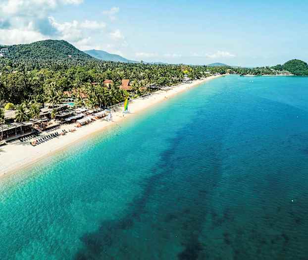 En sandstrand med turkisblått hav, omgitt av trær og hus, med folk som slapper av. (AI-generert tekst)