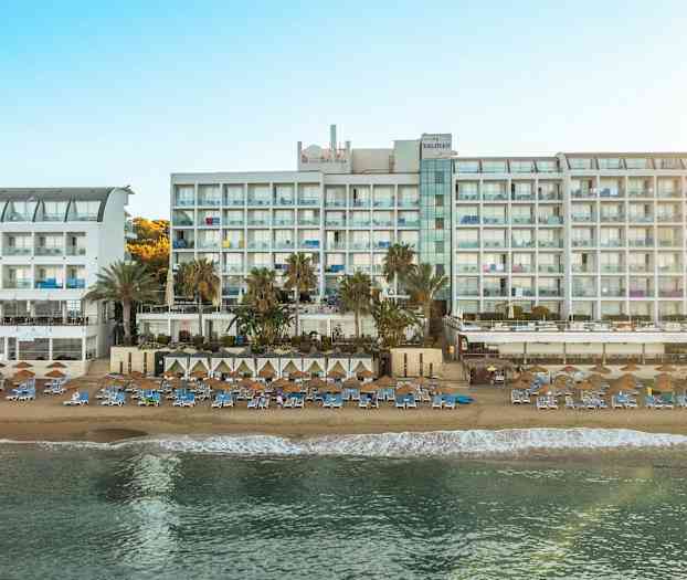 Bilde av en stor hvit hotellbygning ved en solrik strand med solsenger og parasoller. (AI-generert tekst)