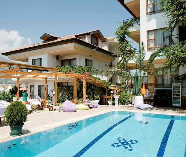 En bygning med hvit fasade, omgitt av grønne planter og med et basseng i forgrunnen. (AI-generert tekst)