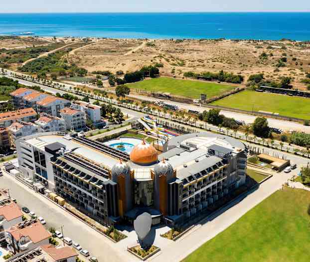 Hotell med unik arkitektur nær havet, omgitt av vegetasjon og grønne områder. (AI-generert tekst)