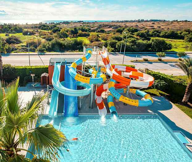 Bildet viser en vannpark med flere fargerike vannsklier og et basseng, omgitt av grøntområder og landskap. (AI-generert tekst)