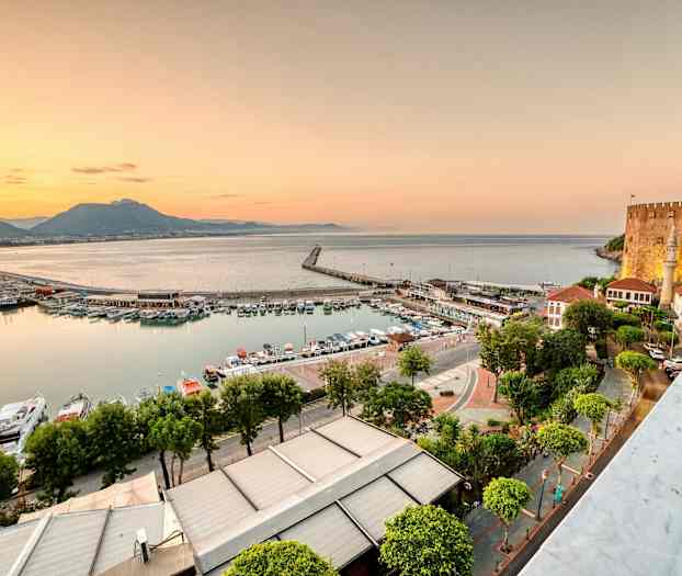 Bildet viser en havn med båter og bygninger ved sjøen under solnedgangen. En borg er synlig på høyre side. (AI-generert tekst)