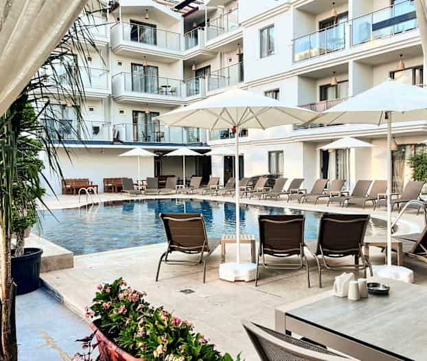 Bildet viser et basseng ved et hotell omgitt av solsenger, parasoller og balkonger i bakgrunnen. (AI-generert tekst)