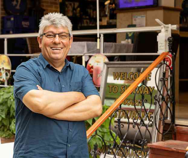 En person står med armene i kryss foran en restaurantinngang med en velkomstskilt. (AI-generert tekst)