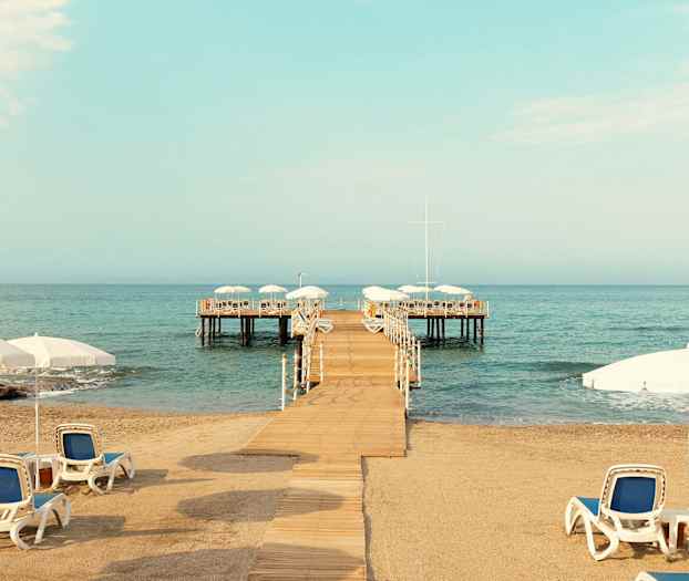 Sandstrand med solsenger og parasoller, en brygge som leder ut til en plattform over vannet. (AI-generert tekst)