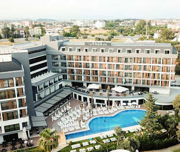 Flyfoto av hotellbygning med bassengområde og solsenger foran. (AI-generert tekst)