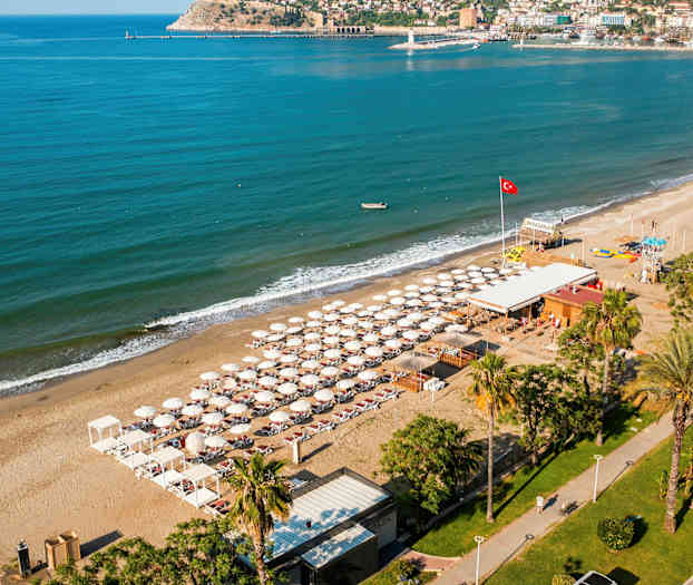 Luftfoto av en strand med solsenger og parasoller, omgitt av turkis hav og bygninger i bakgrunnen. (AI-generert tekst)