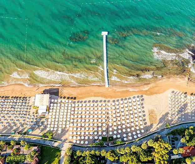 Et flyfoto av en strand med parasoller og solsenger samt en brygge som strekker seg ut i turkis vann. (AI-generert tekst)