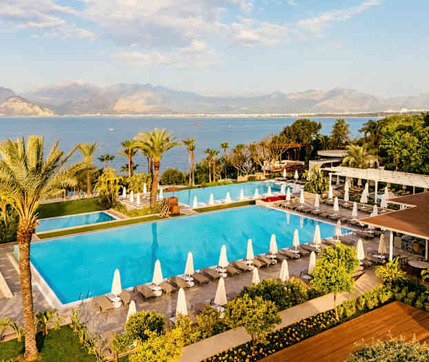 Bilde av et stort basseng omgitt av solsenger med parasoller, grøntområder og utsikt over havet og fjellene i bakgrunnen. (AI-generert tekst)