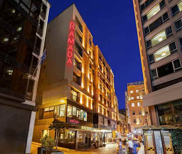 Bildet viser fasaden til et hotell med teksten "RAMADA" opplyst i rødt, omgitt av andre bygninger og en kveldsopplyst bymiljø. (AI-generert tekst)