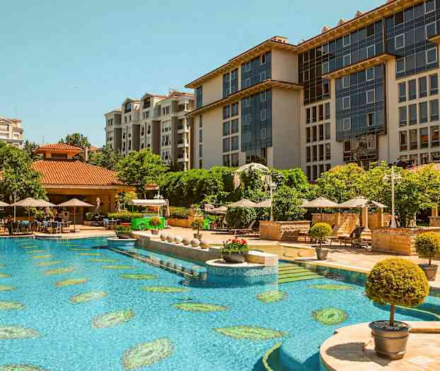 Hotellområde med et stort basseng, solsenger og parasoller omgitt av grønne trær og moderne arkitektur. (AI-generert tekst)