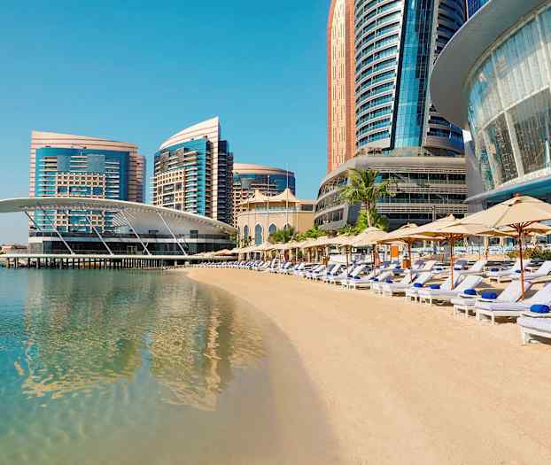 En strand med solsenger og parasoller foran moderne høyhus ved sjøen. (AI-generert tekst)