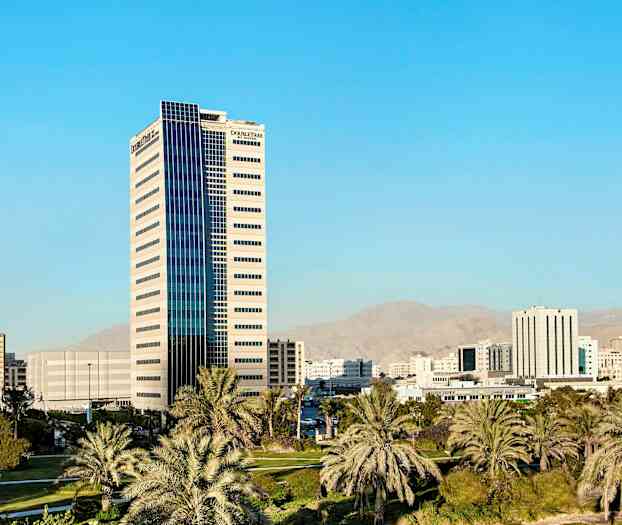 En byutsikt med en høy bygning omgitt av palmer og andre bygninger i bakgrunnen. (AI-generert tekst)