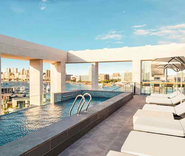 En takterrasse med basseng har utsikt over en by og havn, omgitt av solsenger og parasoller. (AI-generert tekst)