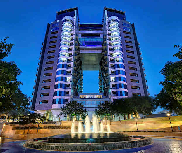 En moderne bygning med to tårn sammenkoblet med en bro. I forgrunnen er en fontene omgitt av trær. (AI-generert tekst)