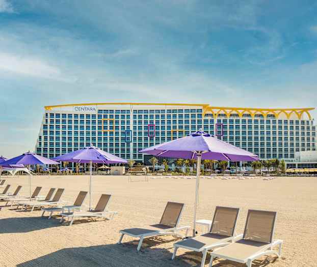 Bildet viser en sandstrand med solsenger og lilla parasoller. I bakgrunnen er en stor bygning med flere etasjer. (AI-generert tekst)