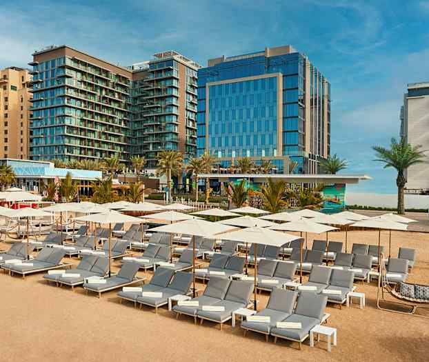 Solstoler og parasoller på sandstrand foran hotellbygninger. (AI-generert tekst)