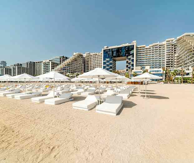 Solstoler og parasoller på en sandstrand med et hotellkompleks i bakgrunnen. (AI-generert tekst)