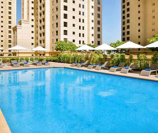 Et basseng med solsenger og parasoller omgitt av høye bygninger. (AI-generert tekst)