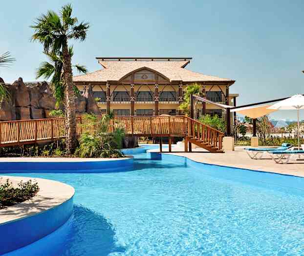 Et svømmebasseng med blått vann og en trebro, omgitt av palmer og bygninger. (AI-generert tekst)