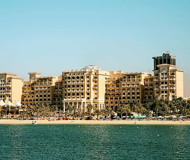 En stor hotellbygning ved en strand med palmer foran, en lys himmel i bakgrunnen og havet i forgrunnen. (AI-generert tekst)