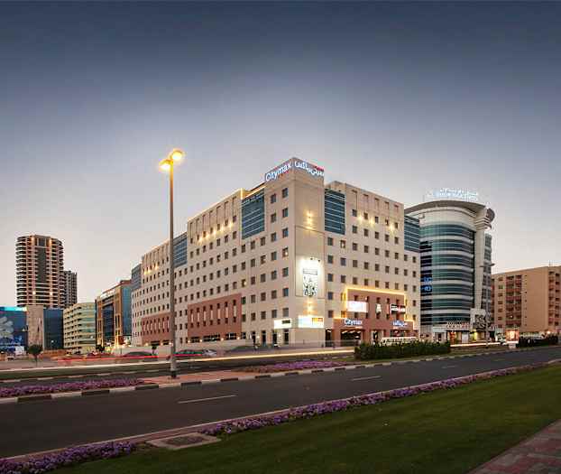 Bymiljø i skumringen med leilighetsbygg og moderne hotellbygg. En vei med biltrafikk og blomster langs fortauet. (AI-generert tekst)