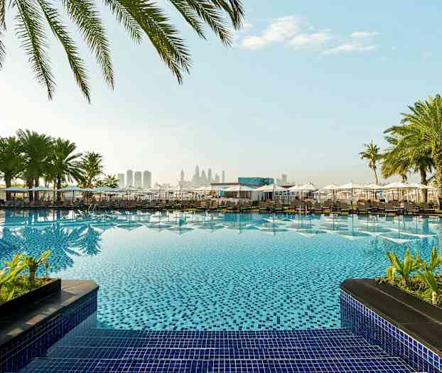 Hotellbasseng omgitt av parasoller og palmer med bysilhuett i bakgrunnen. (AI-generert tekst)