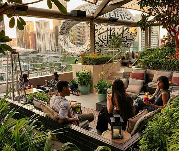 En utendørslounge med sitteplasser omgitt av grøntområder, med en byutsikt og en moderne bygning i bakgrunnen. (AI-generert tekst)