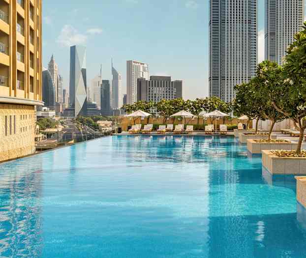 Et infinitybasseng omgitt av solsenger og parasoller, med høye bygninger i bakgrunnen. (AI-generert tekst)