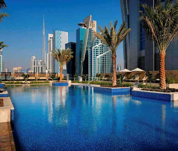 Et svømmebasseng omgitt av palmer med moderne bygninger i bakgrunnen. (AI-generert tekst)