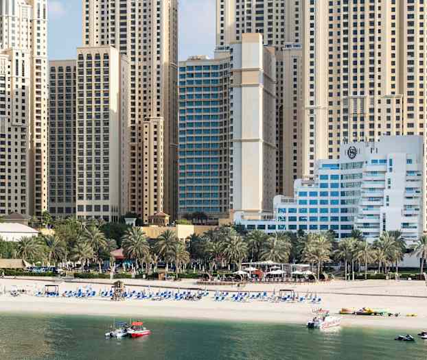 Høye bygninger i bakgrunnen med en strand foran samt palmer. (AI-generert tekst)