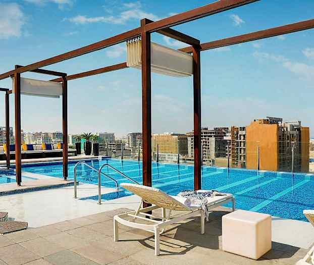 Et utendørsbasseng med utsikt over en by. Solsenger, bord og dekorasjoner er plassert rundt bassenget. (AI-generert tekst)