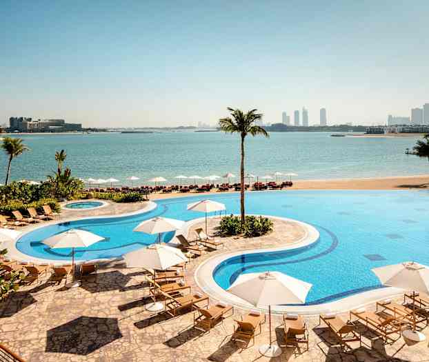 Et hotellbasseng med solsenger og parasoller plassert ved en strand med utsikt over havet og en by i bakgrunnen. (AI-generert tekst)