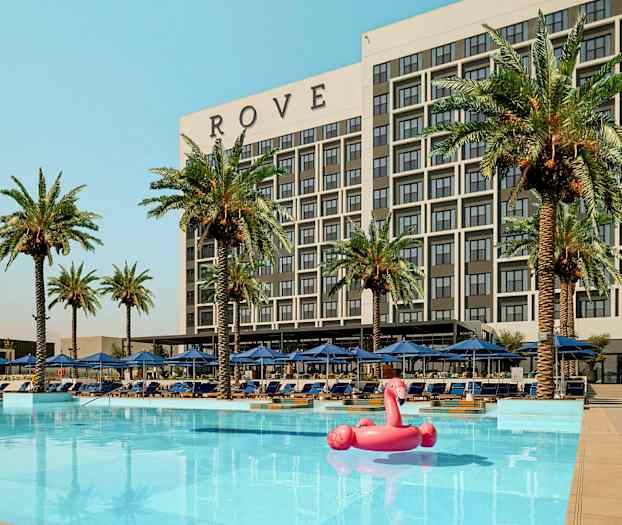 En bygning merket 'ROVE' bak et basseng med palmer og solsenger rundt og en rosa flamingo oppblåsbar leke i vannet. (AI-generert tekst)