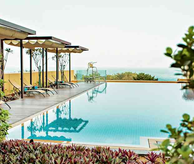 Et bilde av et utendørs svømmebasseng med solsenger under skyggefulle tak, omgitt av vegetasjon og hav i bakgrunnen. (AI-generert tekst)