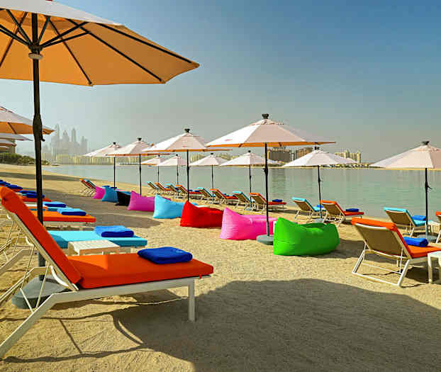 En sandstrand med solsenger med fargerike puter og parasoller ved et rolig vann. (AI-generert tekst)