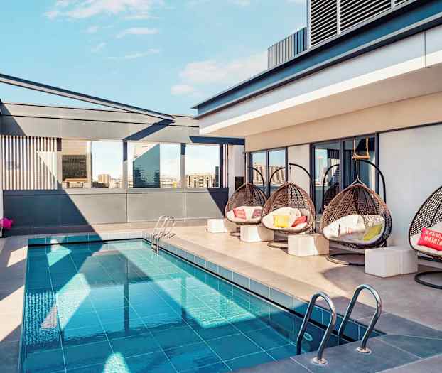 En takterrasse med et lite basseng, hengende flettede stoler og moderne omgivelser. Bygninger er synlige i bakgrunnen. (AI-generert tekst)