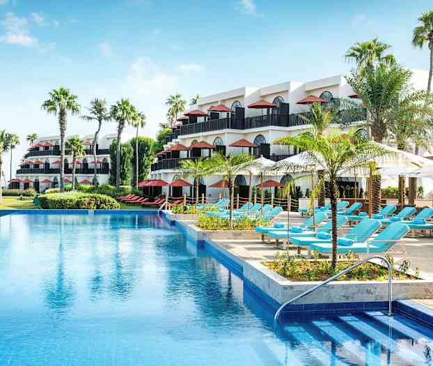 Et hotellområde med flere etasjer, palmer, et svømmebasseng og solsenger under parasoller. (AI-generert tekst)