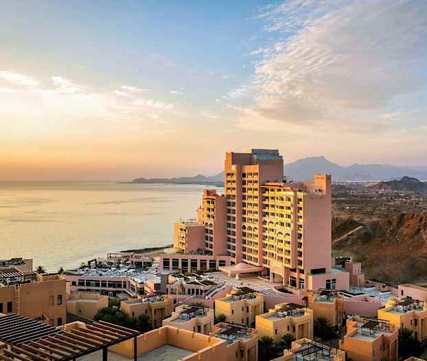 Hotellbygning nær havet ved solnedgang, omgitt av mindre bygninger og fjell i bakgrunnen. (AI-generert tekst)