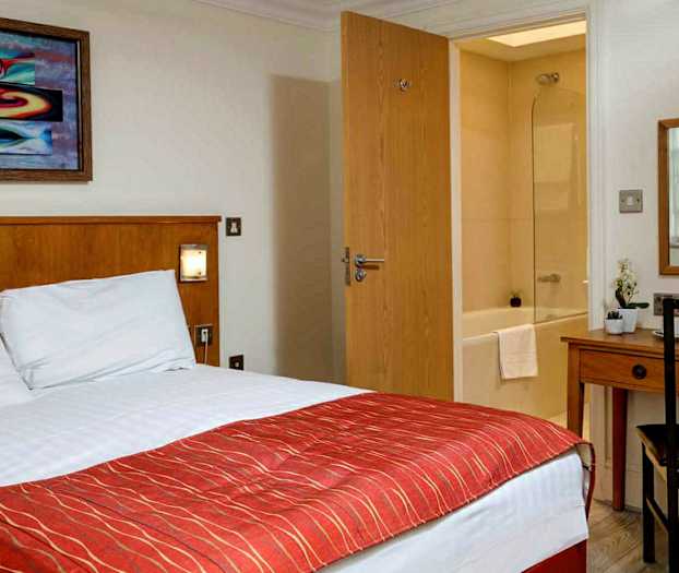 Hotellrom med seng, skrivebord, speil og utsikt mot et bad. (AI-generert tekst)