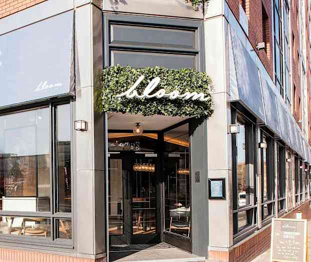 Inngangen til restauranten Bloom med store vinduer og et skilt med navnet omgitt av grønt. (AI-generert tekst)
